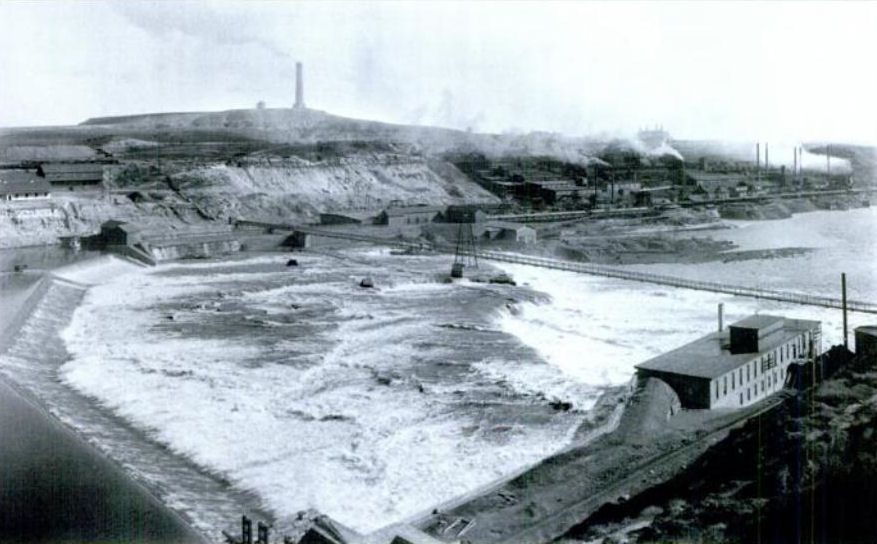 Black Eagle Dam on the Missouri River with smelter buildings and a smokestack on the bluff, before 1908
